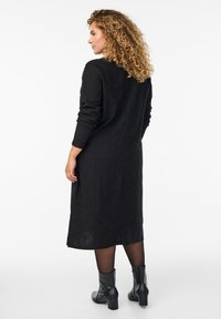 Žena s kovrčavom kosom u dugoj crnoj teksturiranoj haljini, crnim tajicama i crnim čizmama do gležnjeva, stoji i gleda sa strane.