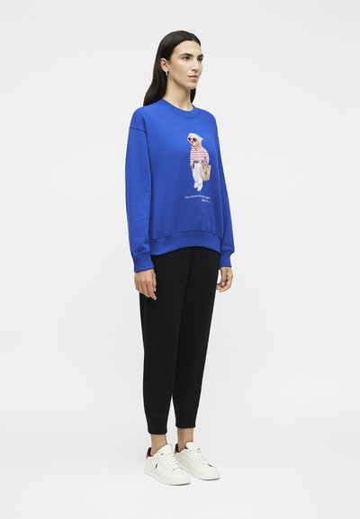 Γυναίκα που φοράει μπλε φούτερ με σχέδιο αρκούδας, μαύρο παντελόνι και λευκά αθλητικά, στέκεται σε λευκό φόντο.