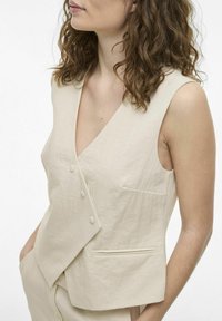 Beige liivi syvällä V-aukolla, jossa on kolme etupainiketta ja sivutasku. Valmistettu teksturoidusta kankaasta, suunniteltu ilman hihoja.