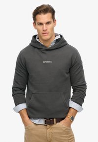 Sudadera gris oscuro con bolsillo frontal, puños acanalados y logotipo bordado "SUPERDRY". Llevada sobre una camisa de rayas azul claro y blanca.