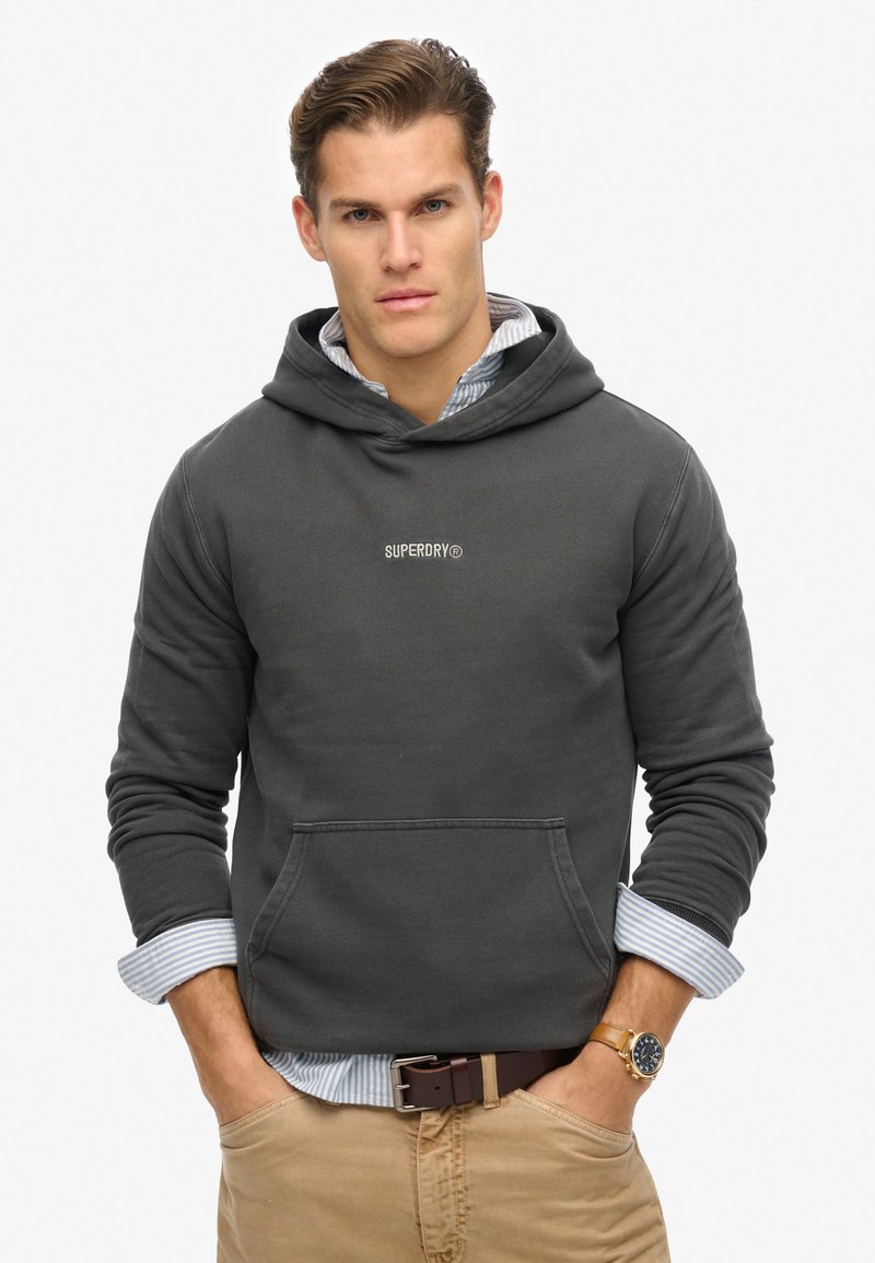 Sudadera gris oscuro con bolsillo frontal, puños acanalados y logotipo bordado "SUPERDRY". Llevada sobre una camisa de rayas azul claro y blanca.