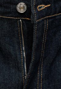 Temno modra denim tkanina z vidnim kovinskim zadrgo in srebrnim gumbom, ki ima kontrastno oranžno barvo šivov ter okroglo etiketo.