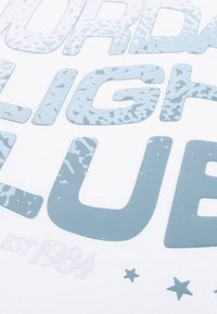 Camiseta de tela blanca con texto grande en azul claro con un patrón texturizado, que dice "SATURDAY LIGHT CLUB" y "EST 1984" con acentos de estrellas.