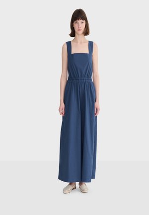 Žena stojaca v bezrukávom tmavomodrom širokých nohaviciach s kockovým výstrihom a elastickým pásom, doplnená béžovými šmykľavými topánkami.