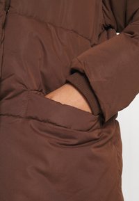 Hnědá zateplená bunda s měkkou texturou, vybavená přední kapsou a manžetami s žebrováním. Tkanina vypadá hladce a lehce.