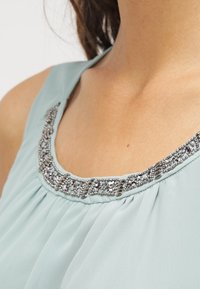 Top senza maniche di colore azzurro chiaro con un design plissettato e un collo decorato con accenti metallici e pietre preziose trasparenti.