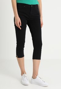 Žena v černých zkrácených úzkých kalhotách s předními kapsami a malými bočními rozparky, v kombinaci s bílými teniskami a zeleným topem.