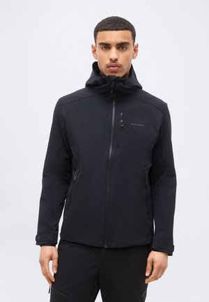 Νεαρός άνδρας φοράει μαύρο μπουφάν με φερμουάρ και κουκούλα, στέκεται μπροστά σε ένα απλό ανοιχτό γκρι φόντο.