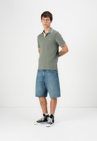 Groene korte mouwen polo met witte randen en een klein logo, gecombineerd met lichtblauwe denim shorts en zwarte hoge sneakers.