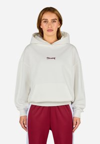 Biely kapucňový sveter s predným vreckom a textom "PEGADOR" v bordovej farbe. Má voľný strih a rebrované manžety. Kombinovaný s červenými nohavicami.