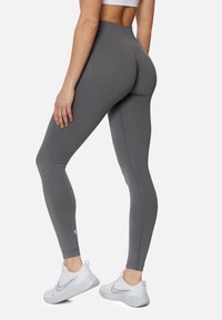 Graue, hoch taillierte Leggings aus glattem, dehnbarem Material. Mit einer figurbetonten Passform und einem subtilen Logodetail nahe dem Knöchel. Getragen mit weißen Sneakers.