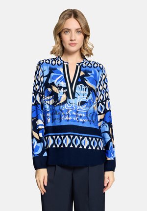 Vrouw die een blauw blouse met geometrische en bloemmotieven draagt, staand met ontspannen handen langs haar zij.