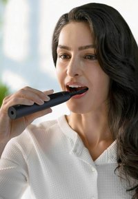 Brosse à dents électrique noire tenue verticalement, avec un design mince. La tête de brosse est positionnée près de la bouche ouverte ; l'utilisateur a des cheveux sombres et ondulés.