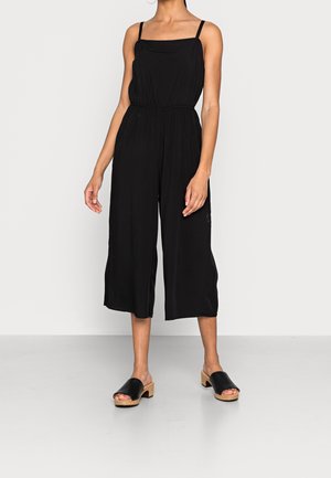 Musta haalaripuku, jossa ohuet olkaimet, joustava vyötärö, leveät lyhyet lahkeet ja sileä kangaspinta. Yhdistetty mustiin puukantaisiin mules-kenkiin.