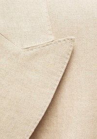 Tessuto beige con motivo a spina di pesce, che mostra una superficie testurizzata e dettagli cuciti su un colletto o un risvolto, enfatizzando il design su misura.