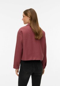 Femme aux longs cheveux bruns portant une veste courte rose poussiére et un jean noir, debout de profil devant un fond uni.