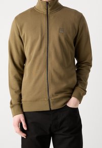 Man med dragkedja upp i olivgrön jacka med hög krage och BOSS-logotyp på, kombinerat med svarta byxor, en hand i fickan.