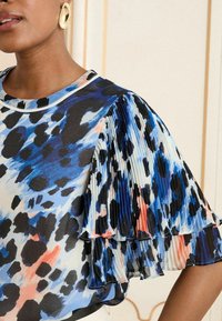 Lühikeste varrukatega topp kergest kangast, millel on sinine, must ja korallne abstraktne trükk. Pleeritud volangid lisavad tekstuuri ja mõõdet.