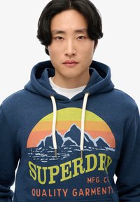 Sudadera color azul marino con un estampado gráfico de montañas y un atardecer en amarillo, naranja y rosa. Presenta dos cordones en color crema y un bolsillo canguro.