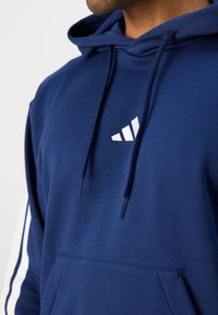 Felpa con cappuccio blu navy realizzata in tessuto morbido, dotata di tasca a marsupio, dettagli bianchi sulle maniche e un logo bianco sul petto.