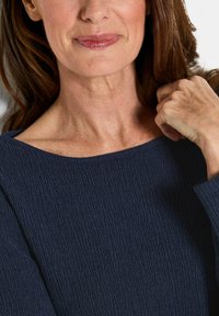 Frau mit braunen Haaren, die ein strukturiertes dunkelblaues Oberteil trägt, sanft lächelnd, mit einer Hand nahe ihrer Schulter.