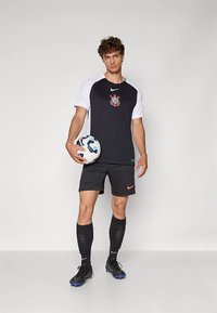 Mees mustvalges jalgpallivormi, hoides valget ja sinist jalgpall, seistes helehallil taustal.