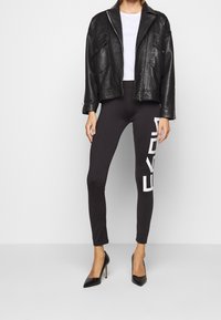 Svart läderjacka med texturerad yta, vit t-shirt, svarta leggings med en stor vit logotyp, och svarta högklackade skor.