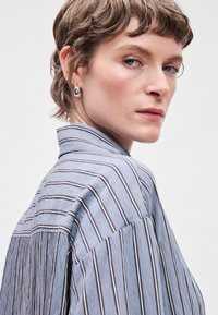 Lyseblå stribet skjorte med opretstående krave og løs pasform. Har en diskret tekstur og vandrette striber, parret med sølvøreringe.