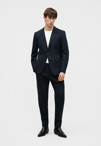 Νεαρός άνδρας με εφαρμοστή μαύρη κοστούμα, λευκό πουκάμισο και μαύρα δερμάτινα παπούτσια, στέκεται μπροστά σε απλό ανοιχτό γκρι φόντο.