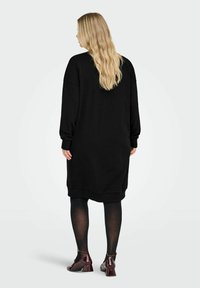 Žena s dugom plavom kosom u labavoj crnoj haljini, crnim tajicama i tamnocrvenim cipelama Mary Jane s niskom petom, stoji okrenuta leđima.