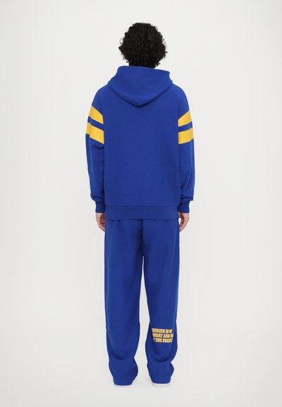 Blaue Sweatshirt mit gelben Streifen auf den Schultern, kombiniert mit passenden blauen Jogginghosen, die unten am Bein gelbe Schriftzüge aufweisen.