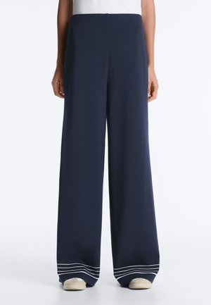 Osoba nosi tmavomodré široké nohavice s bielymi pruhmi pri leme, doplnené bielymi teniskami a bielym topom.