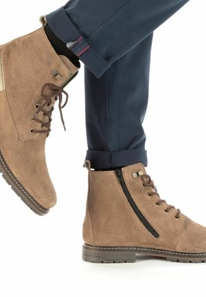Stivali alla caviglia in suede marrone con lacci e chiusura con cerniera, superficie texturizzata e suole in gomma, abbinati a pantaloni blu navy.