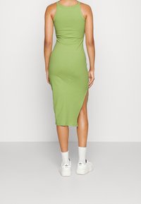 Nainen seisoo selkä kameraan pukeutuneena ilman hihoja, ribbestä kammotessaan vihreään midi-mekkoon, jossa on sivujakso. Hänellä on päällään valkoiset Adidas-tennarit ja sukat yksinkertaisella taustalla.