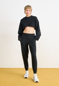 Svart avkortet sweatshirt med lange ermer, kombinert med svarte smale bukser med elastisk midje. Modellen har på seg hvite sneakers.