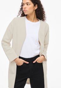 Μπεζ ανοιχτό σακάκι με υφή στην επιφάνεια, μακριά μανίκια και δύο μπροστινές τσέπες, φόρεμένο πάνω από λευκό μπλουζάκι και μαύρο παντελόνι.