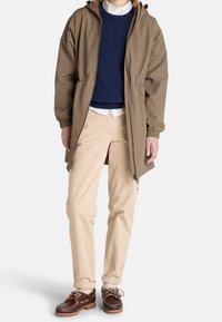 Parka imperméable marron avec capuche à cordon, portée par-dessus un pull bleu et une chemise blanche, associée à un pantalon beige et des chaussures marron à lacets.