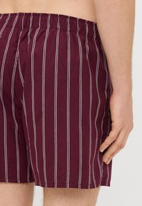 Shorts de baie burgundy cu dungi verticale albe, având un brâu elastic. Materialul pare ușor și are o textură netedă.