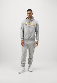Felpa grigia con grafica "Los Angeles Lakers", abbinata a pantaloni jogger coordinati. Entrambi presentano una texture morbida e polsini a coste; scarpe da ginnastica bianche.
