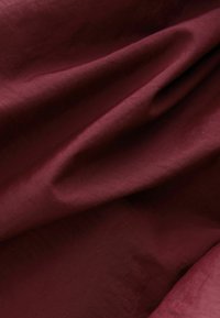 Burgunderroter Stoff mit weicher, glatter Textur, der subtile Falten und ein mattes Finish aufweist und durchgehend eine einheitliche Farbe zeigt.