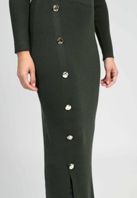 Robe tricotée verte à manches longues, texture côtelée, fente sur le côté et boutons dorés décoratifs le long du côté gauche.