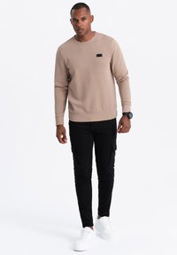 Felpa beige con scollo rotondo e un piccolo etichetta nera, abbinata a pantaloni cargo neri con tasche e sneakers bianche.