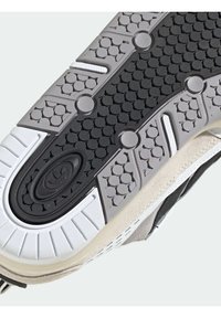 Semelle de chaussure avec un motif de grip en caoutchouc noir, des accents gris et un bord blanc. Comprend un logo au centre et des détails texturés.