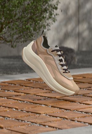 Lysebrun og beige Calvin Klein sneaker med tyk hvid sål, mørke snørebånd, hængende over et trægulv udendørs.