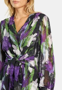 Langes Wickelkleid mit einem floralen Muster in Lila, Grün und Schwarz. Der Stoff ist leicht und semitransparent mit einer gebundenen Taille.