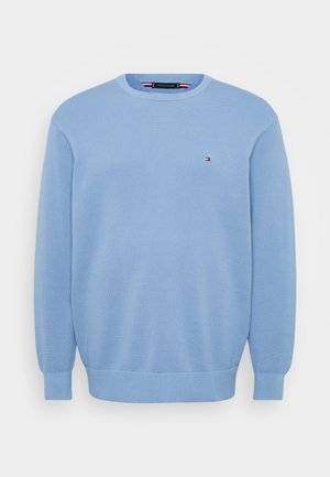 Lyseblå genser laget av strukturert strikket stoff. Har rund halslinning og ribbestrikkede mansjetter, med en liten logo på brystet.