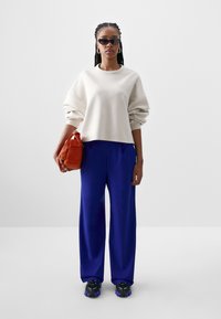 Bijela skraćena dukserica s oversized rukavima, plave široke hlače, crne tenisice s ljubičastim detaljima i narančasta torba.