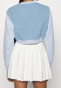 Skórzana bluza z cięciem, w jasnoniebieskim odcieniu z teksturą denimu, z miękkimi niebieskimi rękawami, zestawiona z plisowaną białą spódnicą.