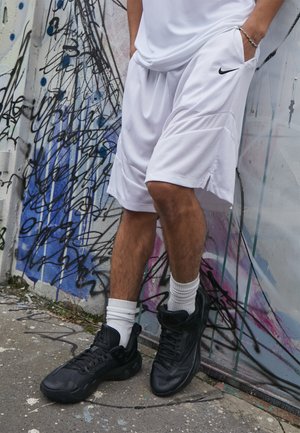 Pantaloni corti bianchi in mesh Nike con tasche laterali, abbinati a scarpe sportive nere. Lo sfondo presenta graffiti colorati su una parete texturizzata.