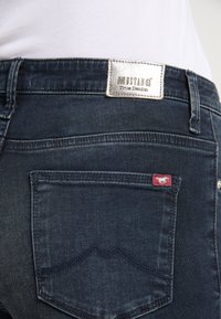 Poche arrière d'un jean Mustang True Denim bleu foncé avec détail cousu et étiquette rouge arborant un logo de cheval blanc.